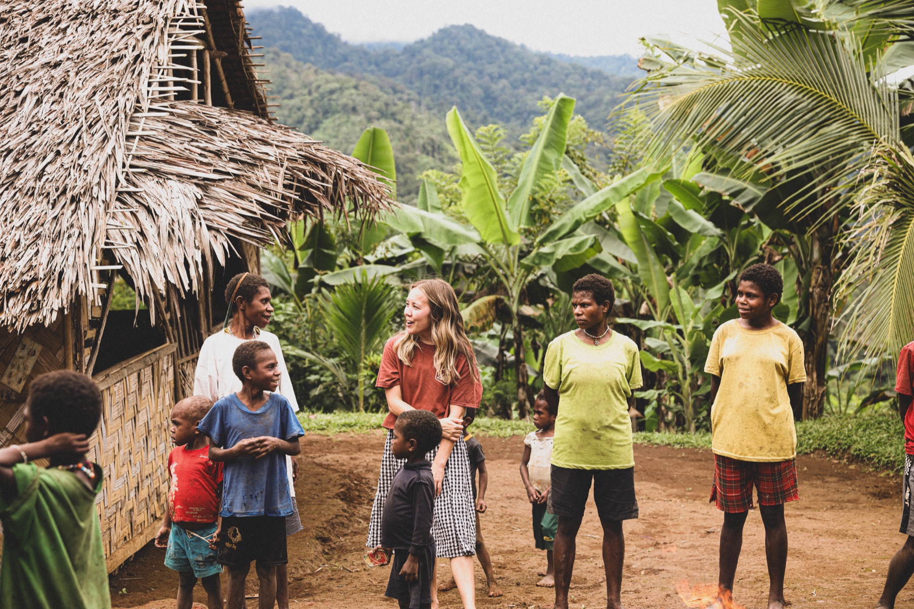 A gathering outside a thatched home, surrounded by lush hills and palms.