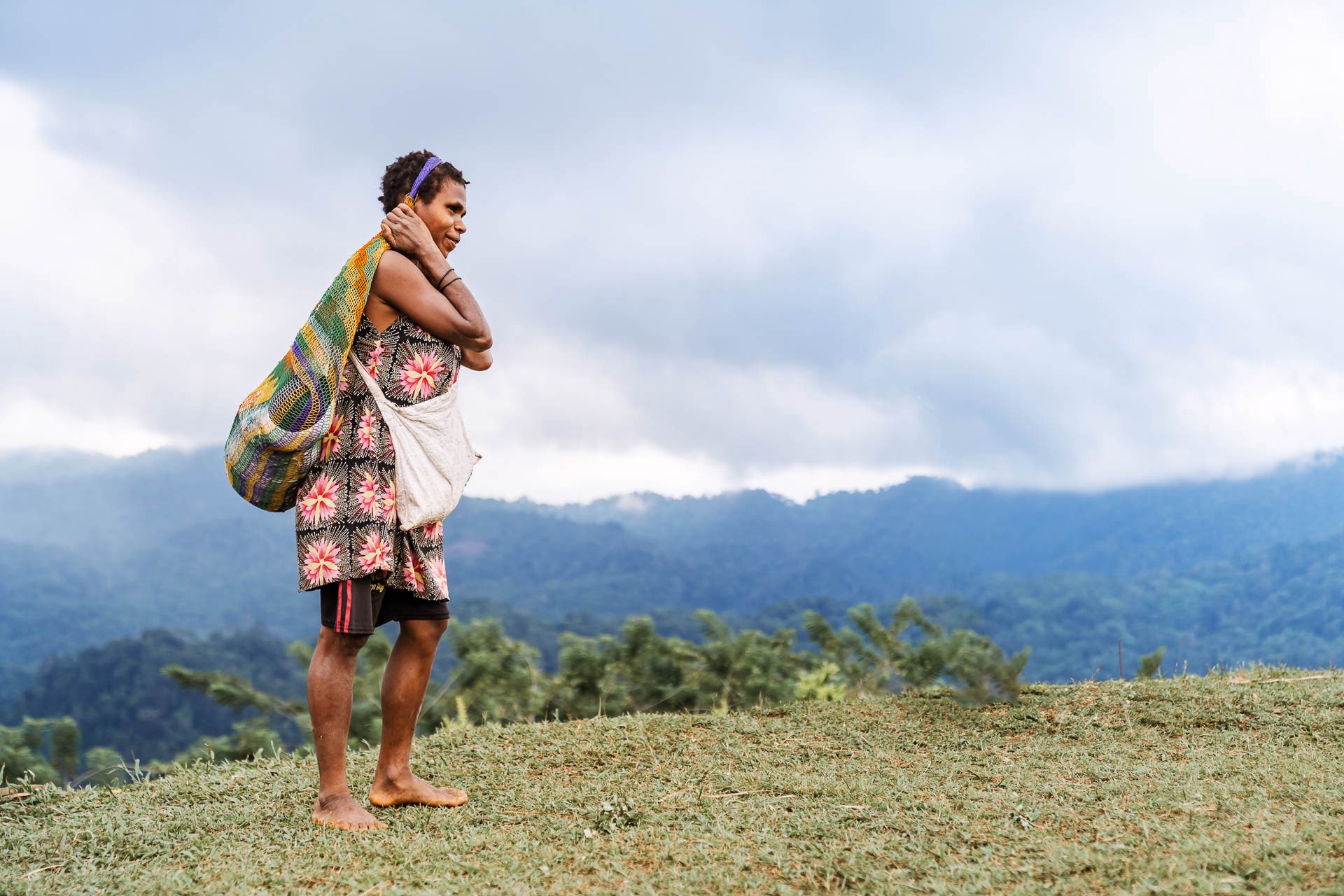 A woman carrying woven bags on a grassy ridge overlooking forested mountains.