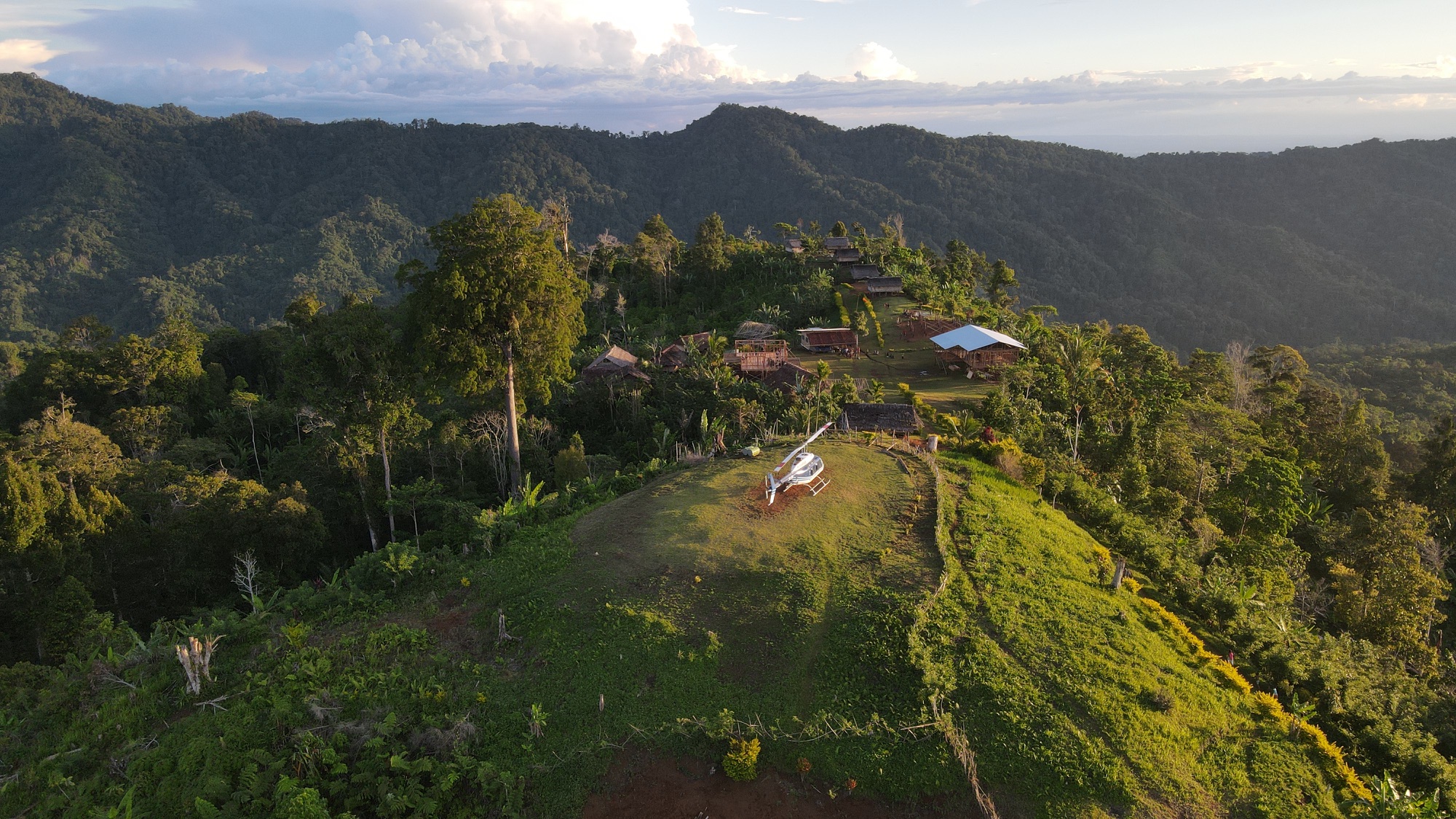 Aerial view of a village along a forested mountain ridge, with a helicopter in a grassy clearing.